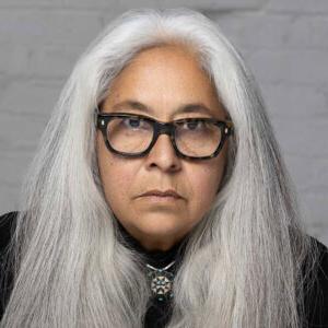 Portrait photo of Laurie Steelink. 斯蒂尔林克有一头又长又直的灰色头发，戴着黑色的上衣和黑框眼镜.
